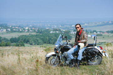 Fototapeta na wymiar Brutal biker with beard wearing leather jacket and sunglasses sitting on his motorcycle on a sunny day, holding helmet. Horizontal picture. Tilt shift lens blur effect