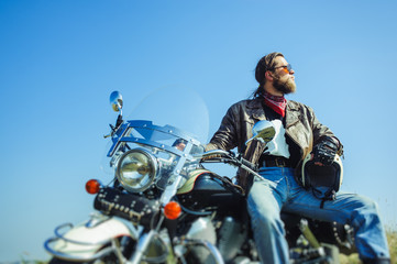 Portrait of a young man with beard sitting on his cruiser motorcycle. Man is wearing leather jacket and blue jeans. Low point of view. Tilt shift lens blur effect