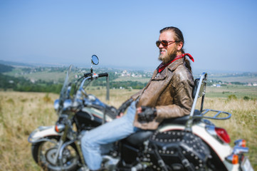 Portrait of biker with long hair and beard in a leather jacket and sunglasses sitting on his bike on the grassy field and smilling. Side view. Tilt shift lens blur effect