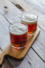 Two glasses of dark amber beer against a wooden rustic cuting board on textured background.
