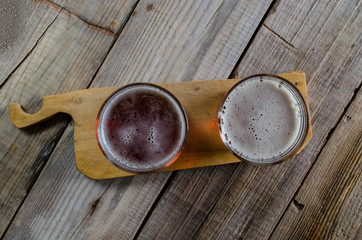 Two glasses of dark amber beer against a wooden rustic cuting board on textured background, top view.