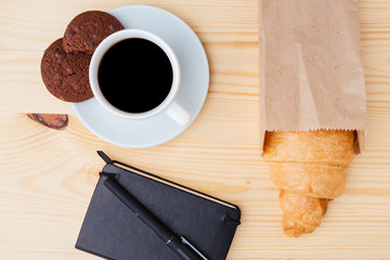 Croissant, coffee and black notebook on wooden table