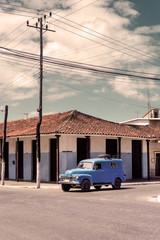 Kuba: Alter blauer US amerikanischer Oldtimer an einer Straßenkreuzung mit typischen Häusern und Strommasten im Hintergrund