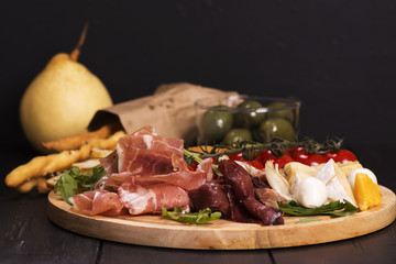 Various types of italian appetizers: ham, cheese, grissini, olives, fruits over black wooden background. Selective focus