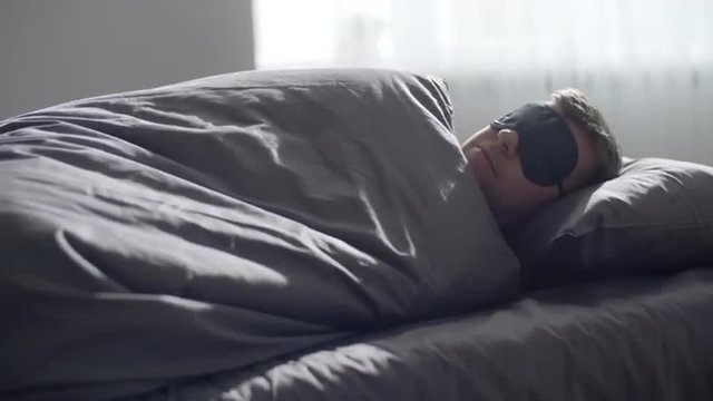 Young man sleeping in eye mask waking up and getting out of bed
