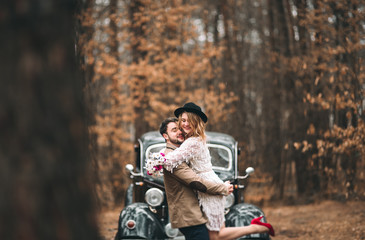 Fototapeta premium Stylish Loving wedding couple kissing and hugging in a pine forest near retro car