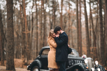 Fototapeta na wymiar Stylish Loving wedding couple kissing and hugging in a pine forest near retro car