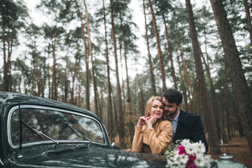 Stylish Loving wedding couple kissing and hugging in a pine forest near retro car