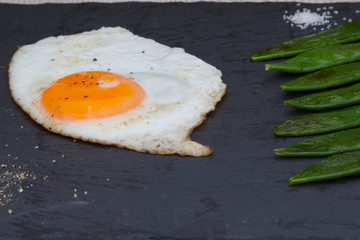Fried egg served with green pea pods on a black stone plate