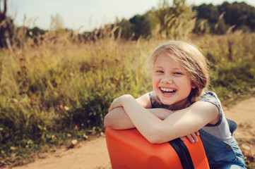happy child girl with orange suitcase travelling on summer vacation. Kid going to countryside. Cozy rural scene