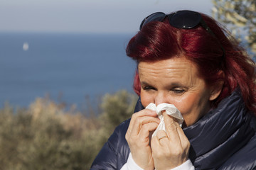 Pretty caucasian middle aged woman with red hair sneezing, blowing nose in handkerchief, outdoors in olive grove above the sea coast, on windy sunny day