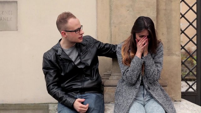 Young, brown hair girl sitting on the stone wall next to the boy in glasses, she is crying and he is cuddling her