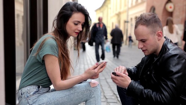 Young brown hair girl sitting on the street and talking with boy. They are  looking at their smartphones and smiling into camera