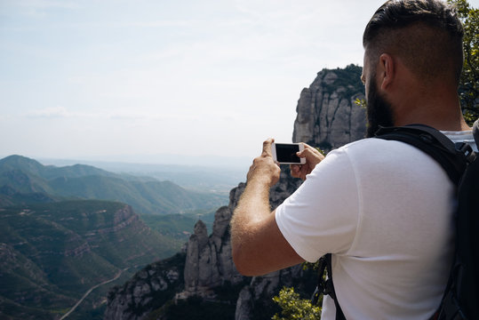 Young male tourist in white t-shirt takes photos with smart phone on peak of rock