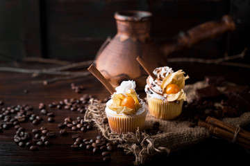 Chocolate cupcakes with white butter cream, decorated with winter cherry on a dark wooden background. Decorated with cinnamon sticks, coffee beans