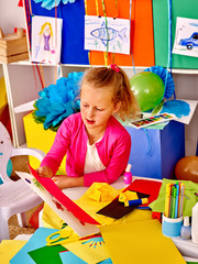 Portrait of female kid keeps colored paper and do origami on table in kindergarten .