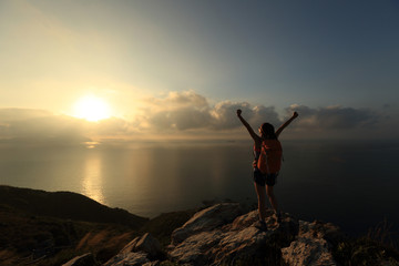 cheering young woman backpacker at sunrise seaside mountain peak