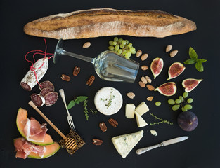 Wine and snack set. Baguette, glass of white, figs, grapes, nuts, cheese variety, meat appetizers, herbs on black grunge background, top view.