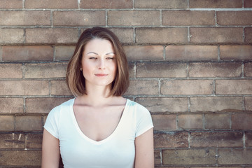 Fototapeta na wymiar Close-up portrait of beautiful innocent Caucasian adult girl woman with long hair, bob style, hazel eyes, in white tshirt standing behind brick wall outside looking away