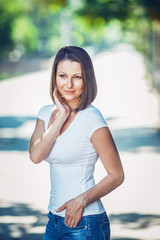 Portrait of beautiful innocent Caucasian adult girl woman with long hair, bob style, hazel eyes, in white tshirt and blue jeans, standing in park street outside looking away from camera