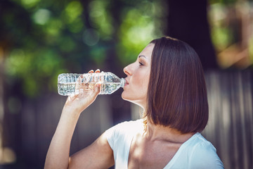 Portrait of beautiful Caucasian adult girl woman with long hair, bob style, in white tshirt drinking clean bottle water in park street outside looking away from camera, profile 