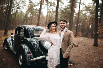 Gorgeous newlywed bride and groom posing in pine forest near retro car in their wedding day