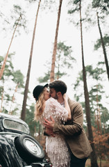 Gorgeous newlywed bride and groom posing in pine forest near retro car in their wedding day