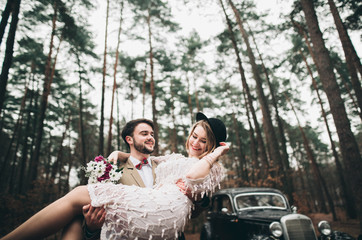 Gorgeous newlywed bride and groom posing in pine forest near retro car in their wedding day