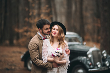 Gorgeous newlywed bride and groom posing in pine forest near retro car in their wedding day
