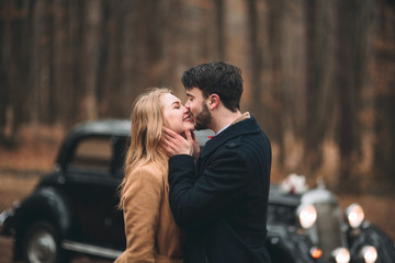 Gorgeous newlywed bride and groom posing in pine forest near retro car in their wedding day