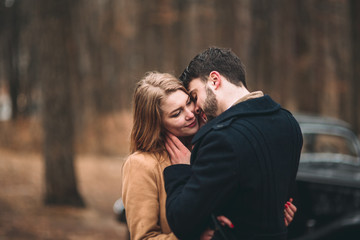 Gorgeous newlywed bride and groom posing in pine forest near retro car in their wedding day