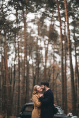 Gorgeous newlywed bride and groom posing in pine forest near retro car in their wedding day