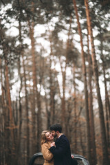 Gorgeous newlywed bride and groom posing in pine forest near retro car in their wedding day