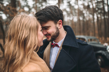 Gorgeous newlywed bride and groom posing in pine forest near retro car in their wedding day