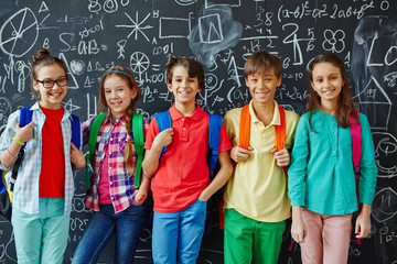 Group of youngsters with backpacks standing against blackboard