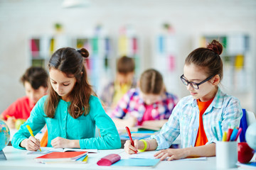 Diligent pupils sitting by desk and writing in copybooks at lesson