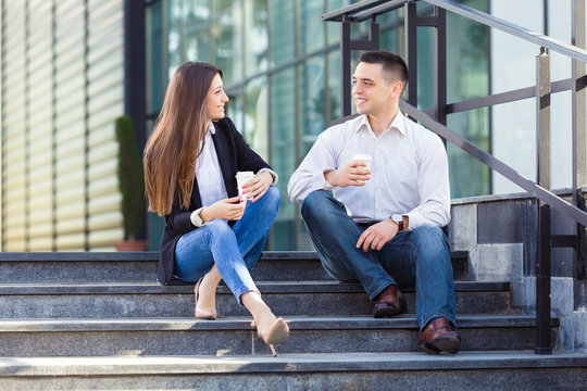 Young businesswoman and businessman sitting on the steps of the office building and drinking coffee
