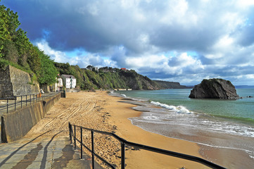 A panorama of the extensive sandy North Beach in Tenby Wales in moody weather with blue sea, a rocky islet and dark dramatic clouds