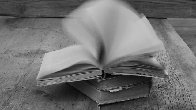 stack of ancient books on old gray wooden boards. strong gusty wind turns the pages of an open book. black and white, monochrome. copy space