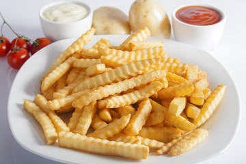 Portion of French fries (Crinkle-cut) deep fried, served on a white plate next to white bowls with mayonnaise and ketchup and fresh potato.