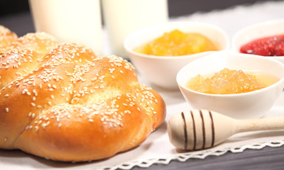 White bowls with homemade jams next to a glass of milk and sweet baked bun are on linen tablecloth on a rustic table