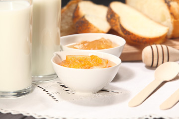 White bowls with homemade jams next to a glass of milk and sweet baked bun are on linen tablecloth on a rustic table