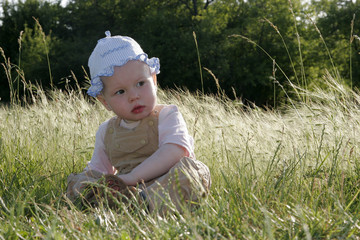 Little girl is sitting on the sunny meadow and looking slightly to the side