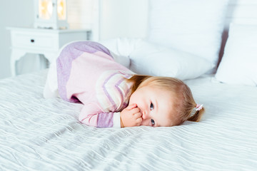 Cute little blonde girl on hands and knees on white bed in bedroom. White interior, big bed, bedside table and night lamp