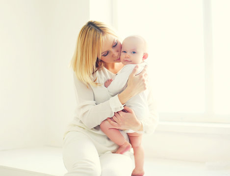 Happy mother kissing baby at home in white room near window