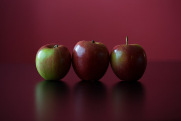 Drei rot-grüne Äpfel, gerade nebeneinanderstehend, vor rotem Hintergrund, mit Spiegelung
