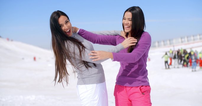 Pair of female friends or sisters laughing while pretending to be strangling each other on ski slope with group of people watching in the background