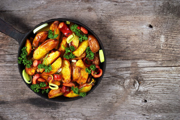 Fried potatoes with sausages, onion rings and green parsley. Meal served in old cast iron frying pan on wooden table.