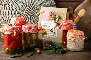 Jars with pickled vegetables and beans, spices, book of recipes and kitchen utensils on wooden background