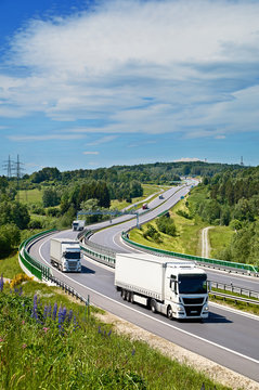 White trucks drive along an asphalt highway with electronic toll gate in a wooded landscape. View from above. Sunny summer day with blue skies and white clouds.